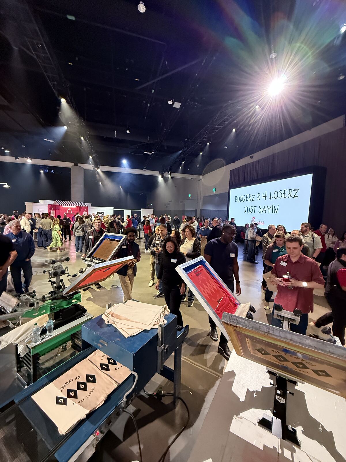 Large indoor event with hundreds of attendees gathered around lit stage displays and merchandise booths at night